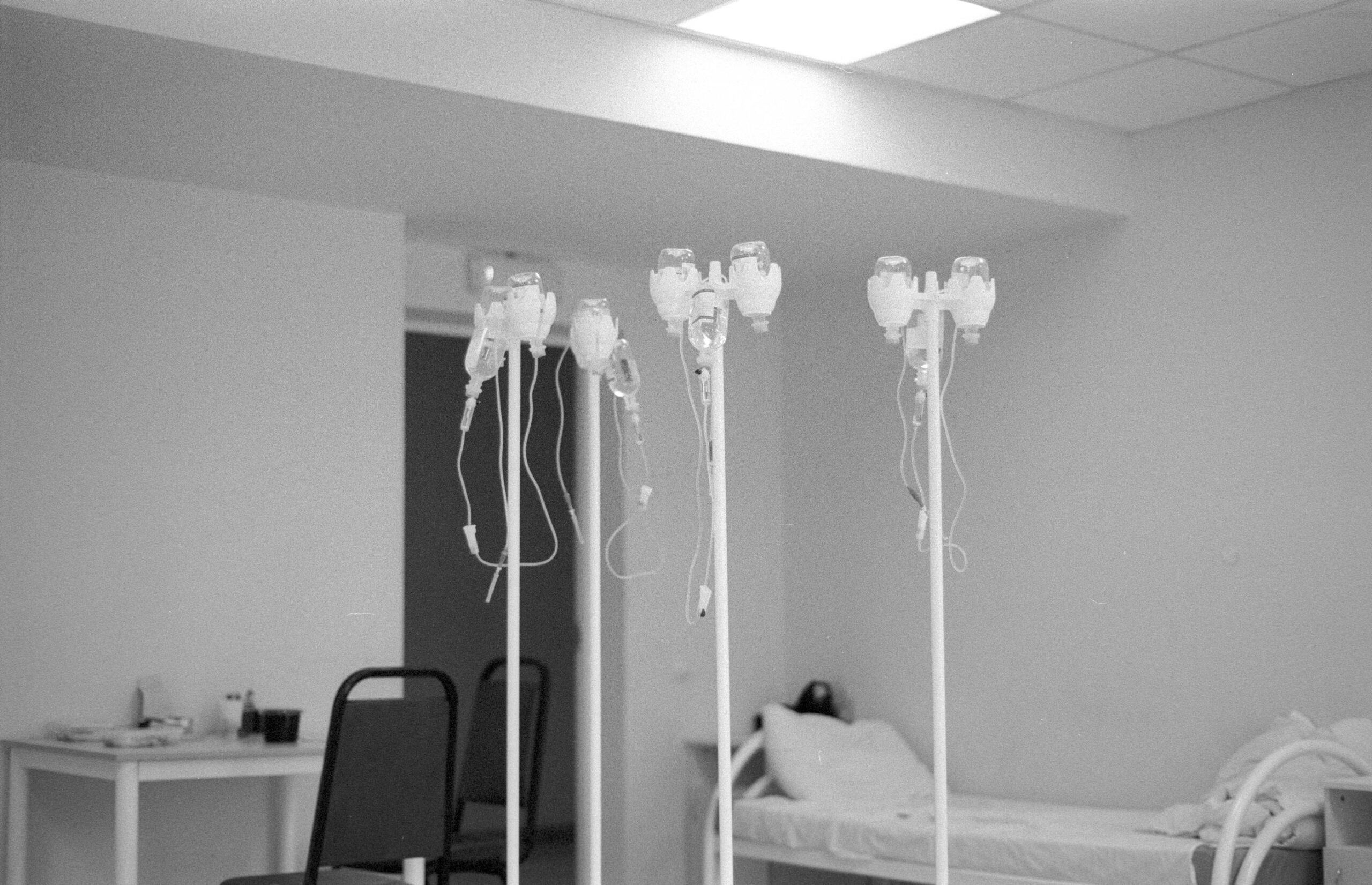 Monochrome photo of a hospital room with IV stands and a bed under fluorescent lights.